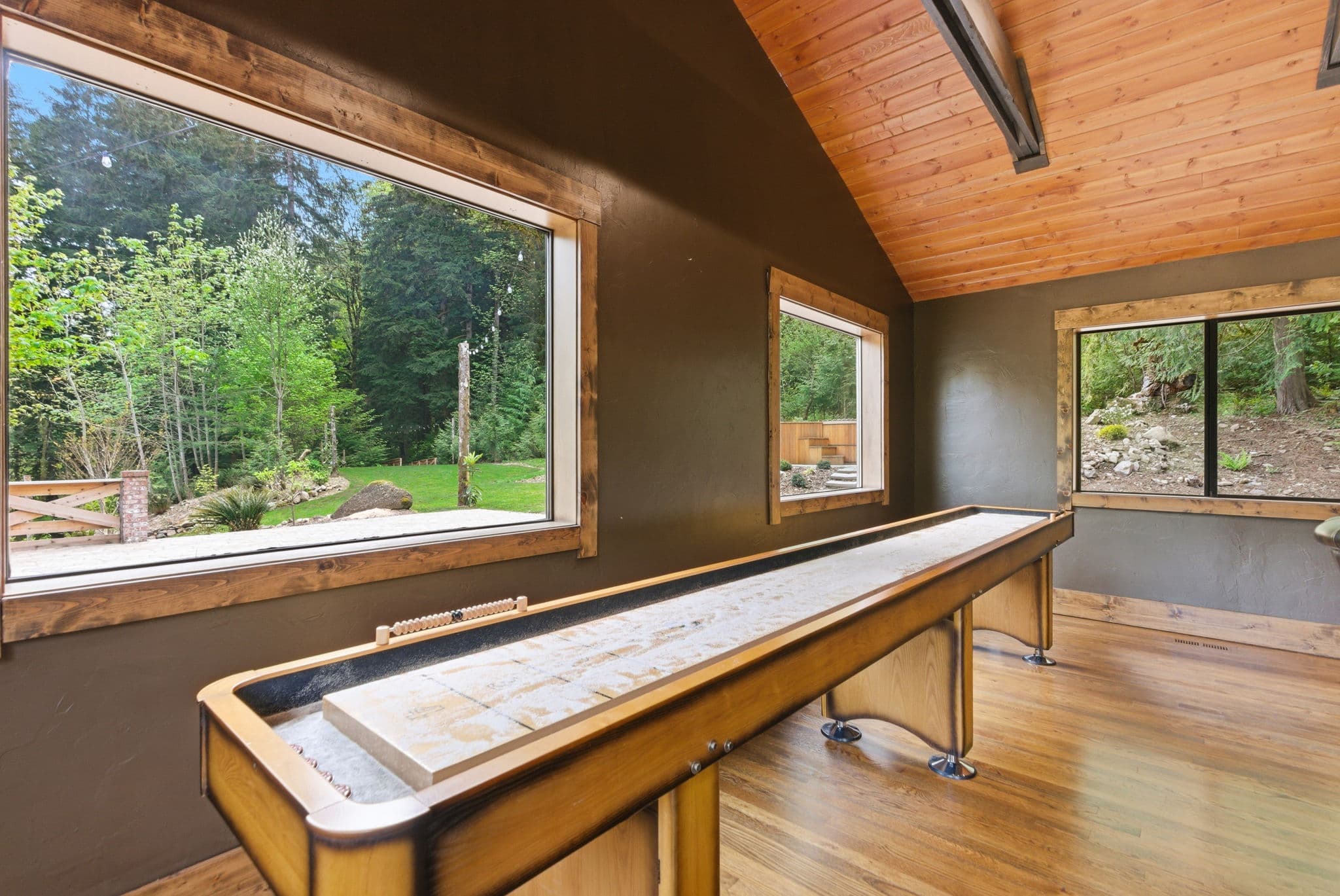 Game room with full-length shuffleboard table and floor-to-ceiling forest windows