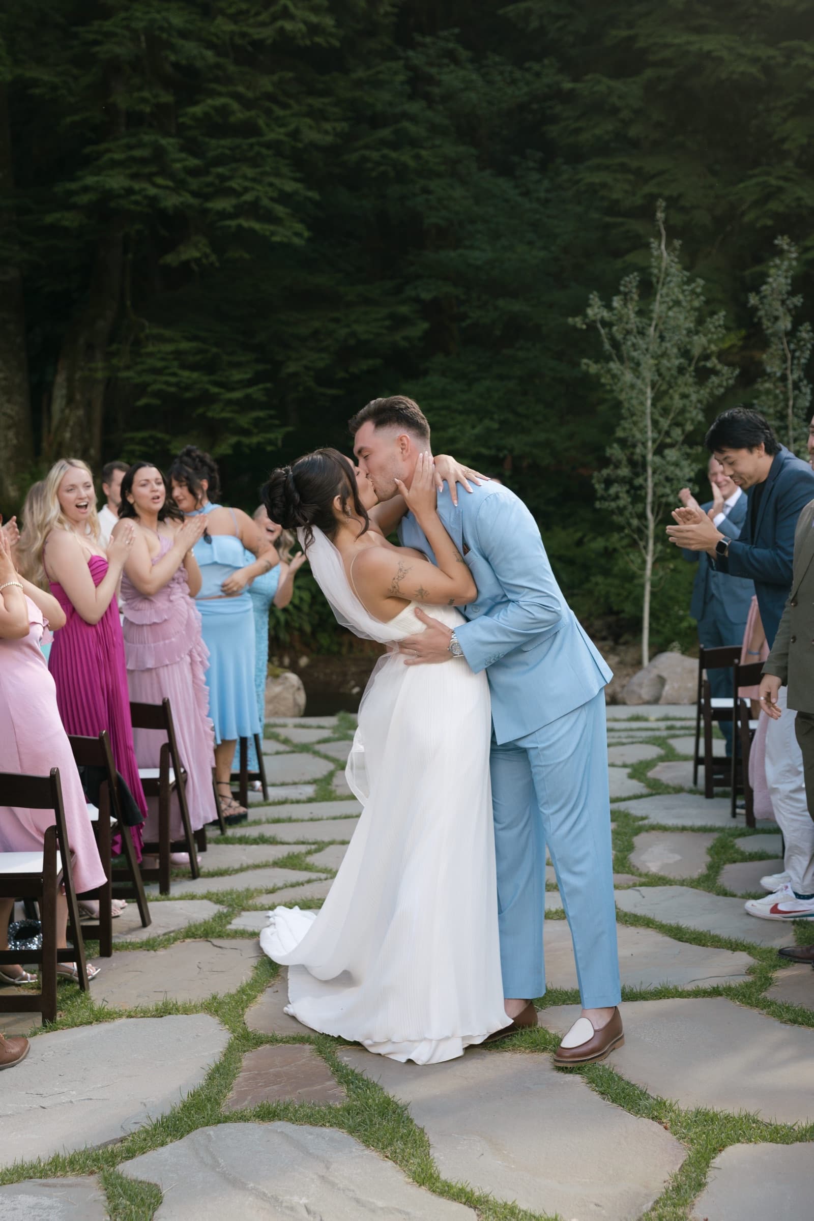 A just-married couple kiss on the flagstone aisle as the bridesmaids cheer