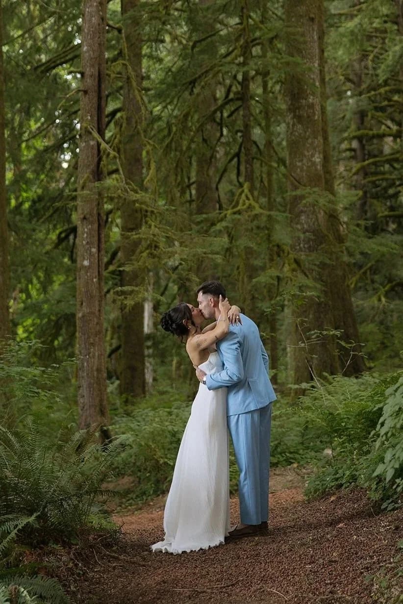 A bride and a groom in a pale blue suit kiss among tall trees